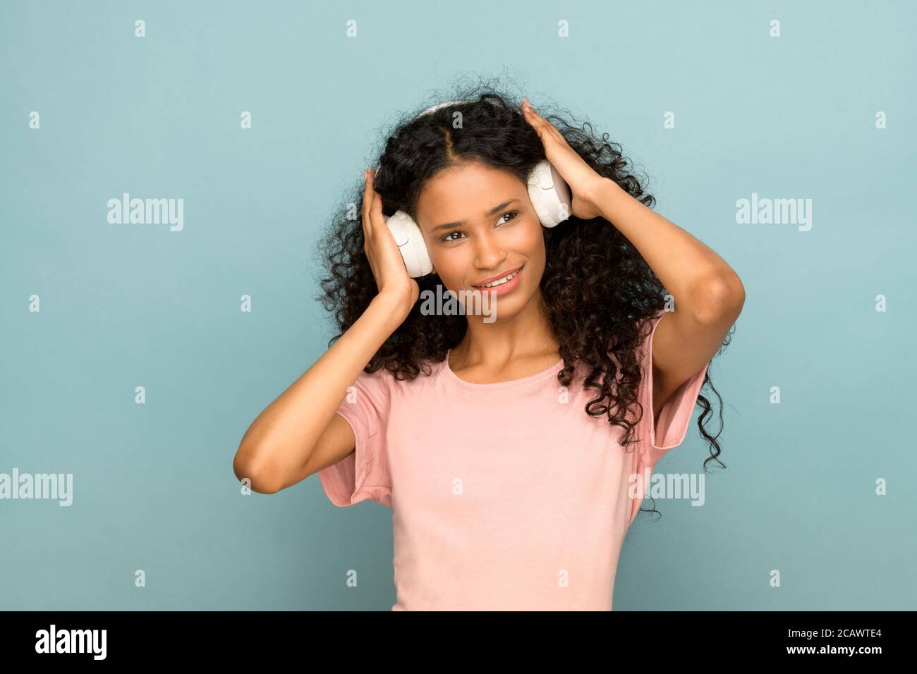 Smiling attractive young Afro American girl with headphones listening to her favorite music in an upper body portrait against a blue studio background Stock Photo