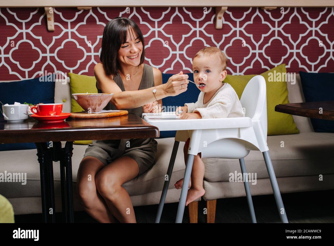 Caring mother in a cafe feeding her baby, sitting in a high chair. Stock Photo