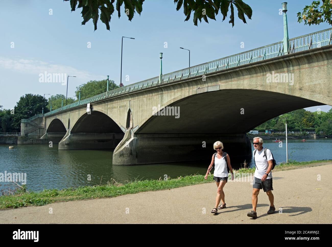 man and woman in shorts and suglasses walk beside the river thames in richmond, surrey, england, with twickenham bridge in background Stock Photo