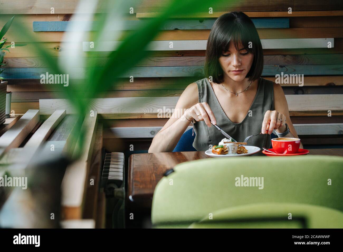 Woman keen on eating cake in a cafe. Frontal shot through a plant. Stock Photo