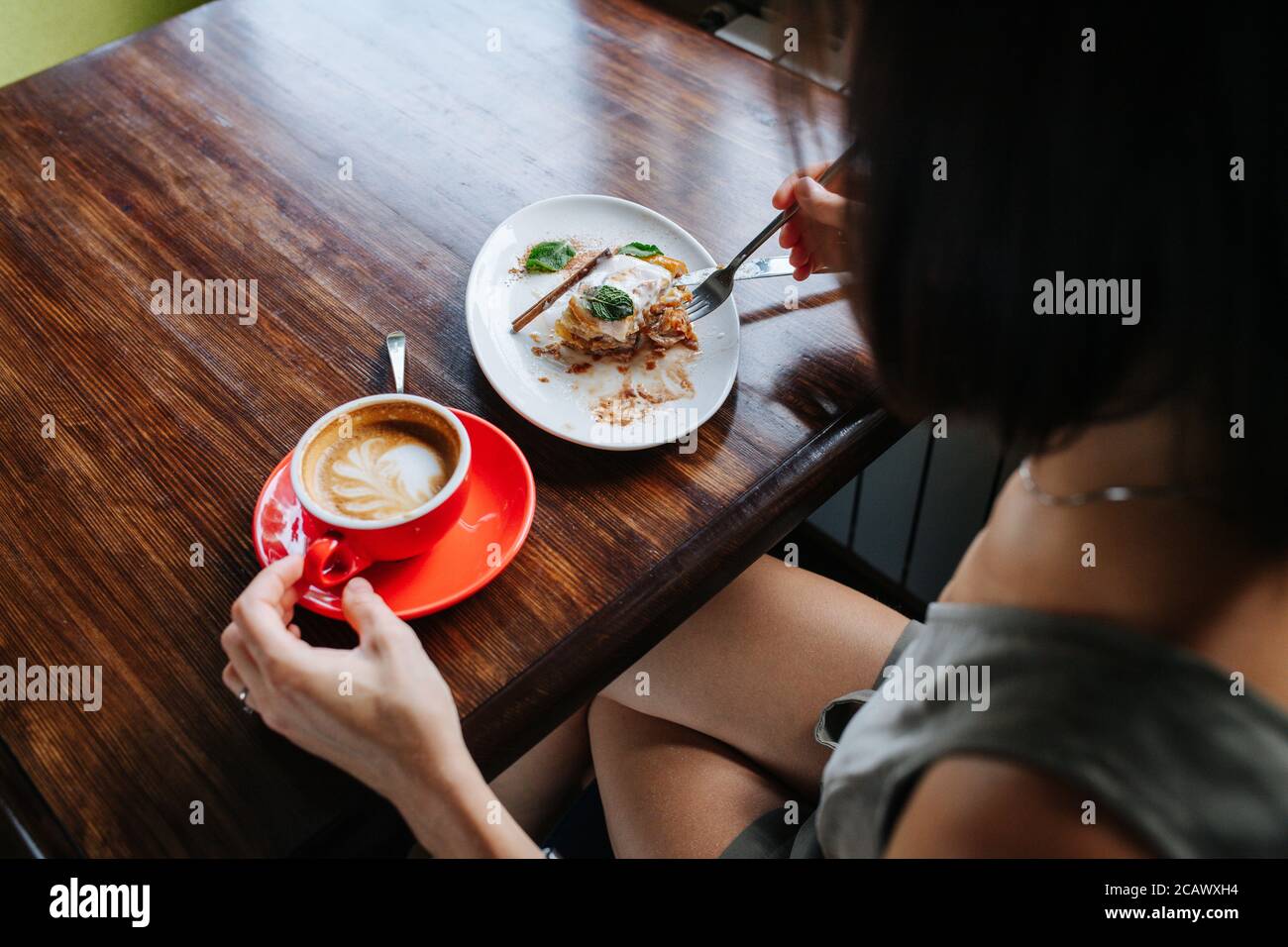 Over a shoulder view of a woman drinking coffee and eating cake in a cafe. Stock Photo