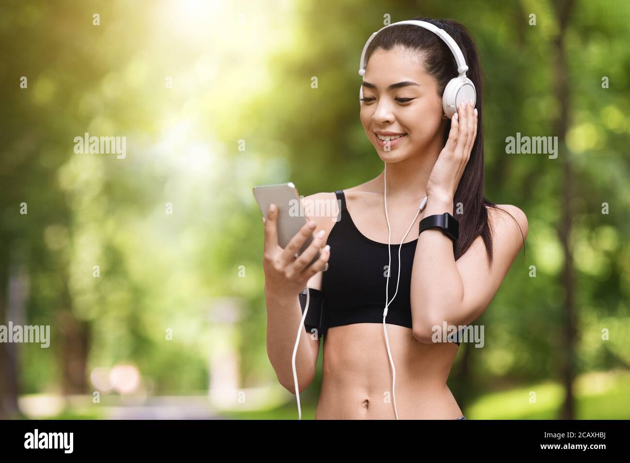 Cheerful Jogger Girl With Smartphone And Headphones Choosing Playlist For Running Outdoors Stock Photo