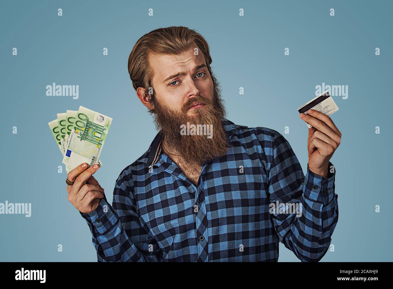 Businessman skeptical choosing holding showing plastic credit card or money cash euro banknotes bills Bearded hipster business man Isolated on blue Ba Stock Photo