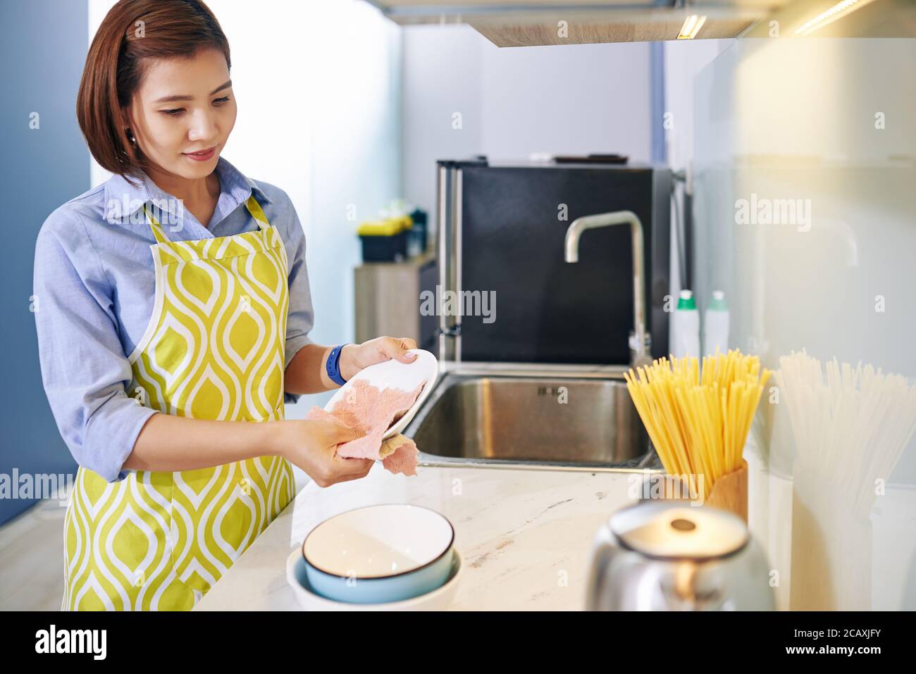 Pretty young Vietnamese housewife wiping plates with soft cloth after finishing cooking Stock Photo