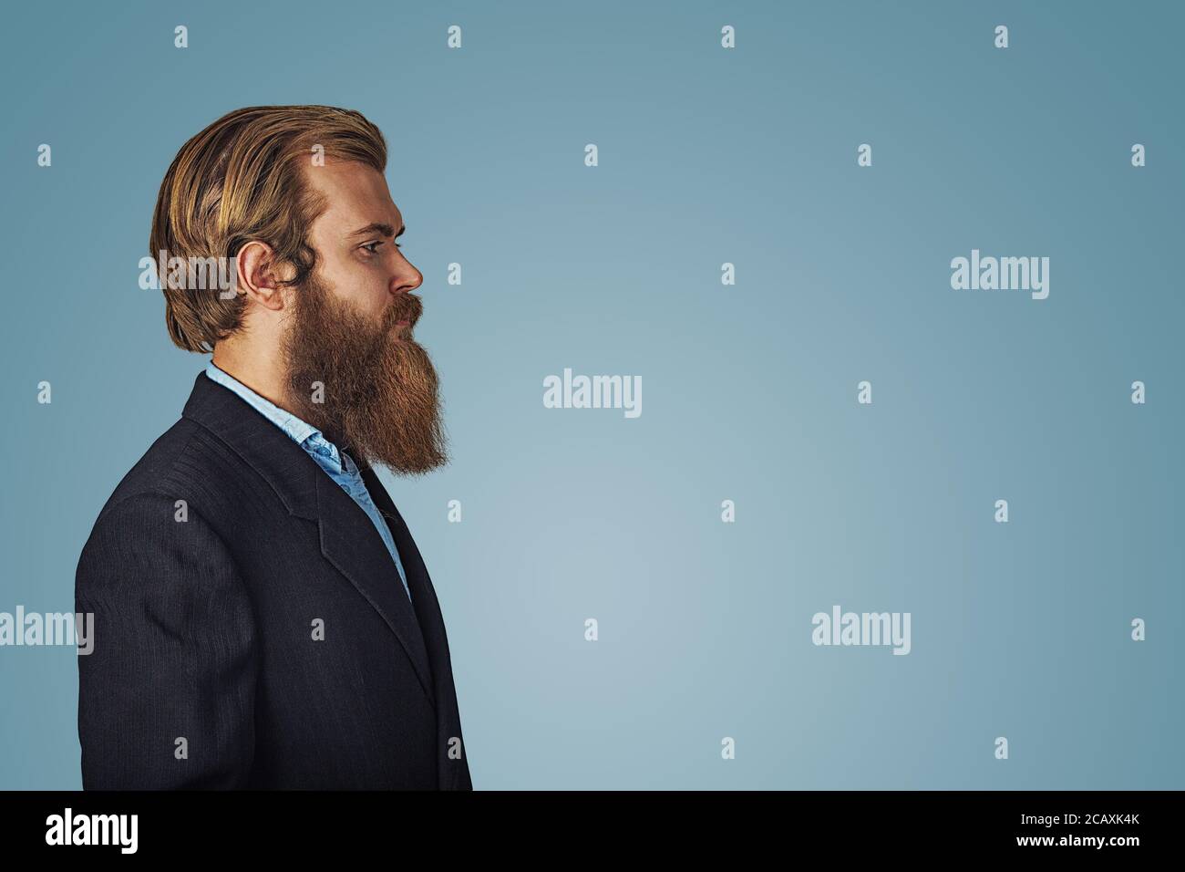 Side view portrait of serious fashionable bearded hipster handsome man in black suit and blue shirt Isolated on blue Background. Negative face express Stock Photo