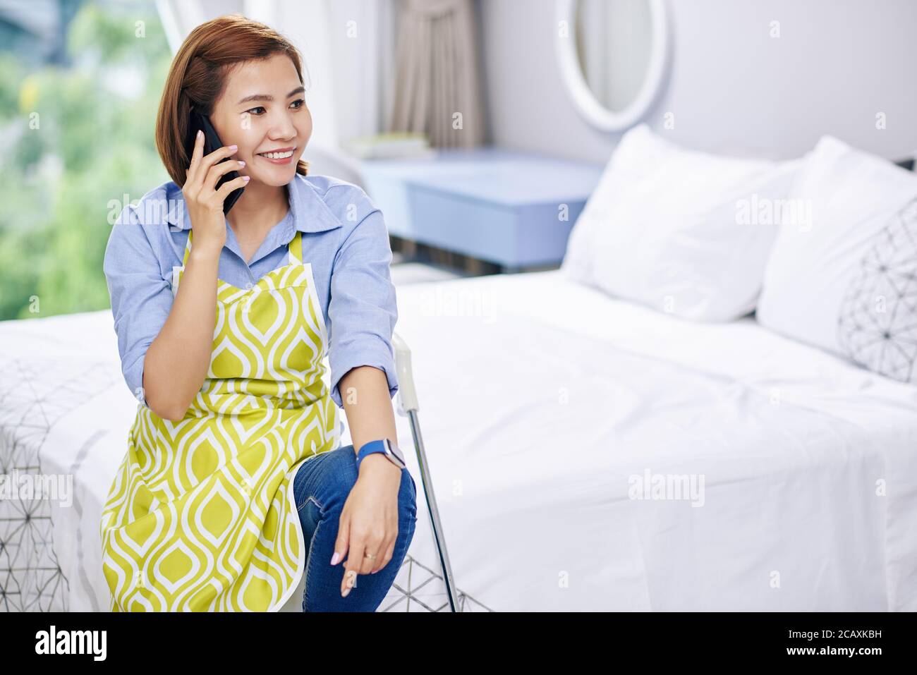 Pretty young Asian housewife talking on phone with friend after cleaning apartment Stock Photo