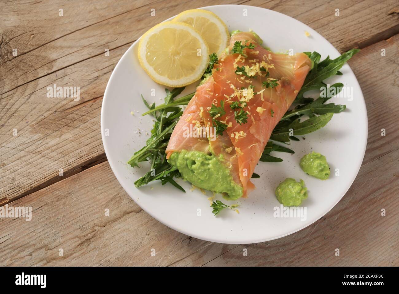 Smoked salmon filled with pea puree on arugula salad with lemon slices and parsley garnish on a rustic wooden table, healthy low carb diet, copy space Stock Photo