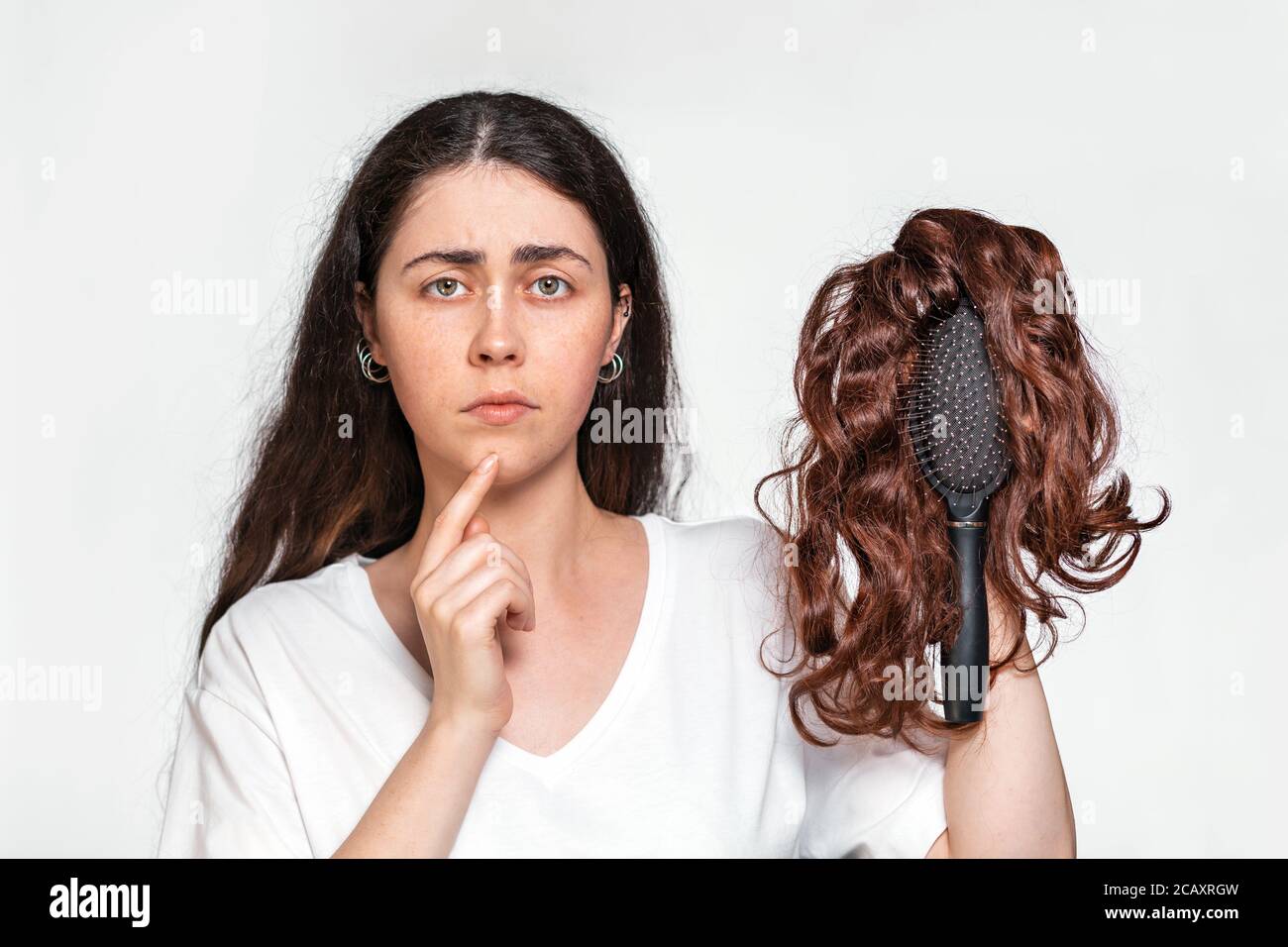 Portrait of a sad woman holds a comb with a wig on it. White background. Concept of hair care and hair loss. Stock Photo