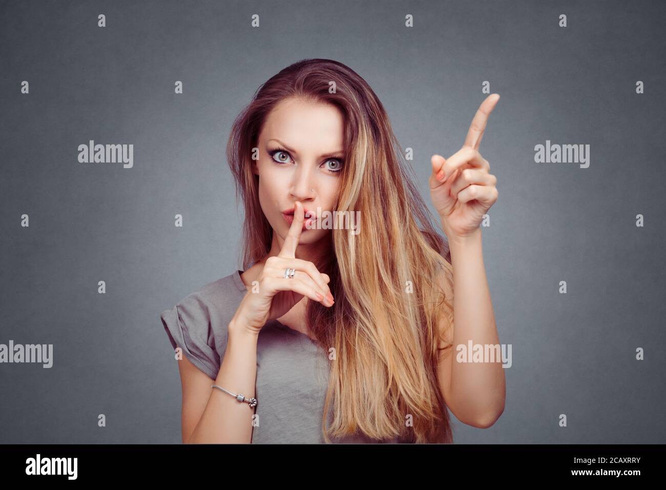 Angry woman showing silence, hush sign gesture with one hand, attention listen to me with other hand isolated on gray grey studio wall background. Neg Stock Photo