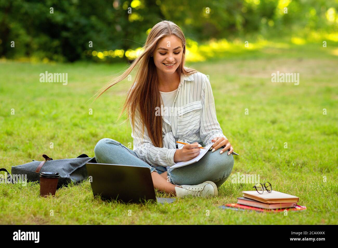 Beautiful student girl preparing for lessons in park, writing notes to workbook Stock Photo