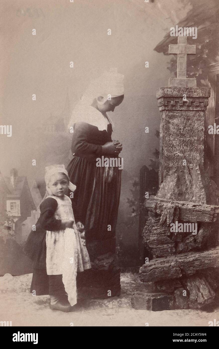 A pregnant woman and child pray in front of cross.  Holland, 1880’s from an old print. Stock Photo