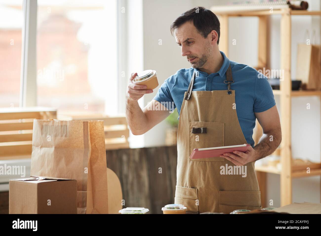 Waist up portrait of mature man wearing apron packaging orders while standing by wooden table, food delivery service worker, copy space Stock Photo