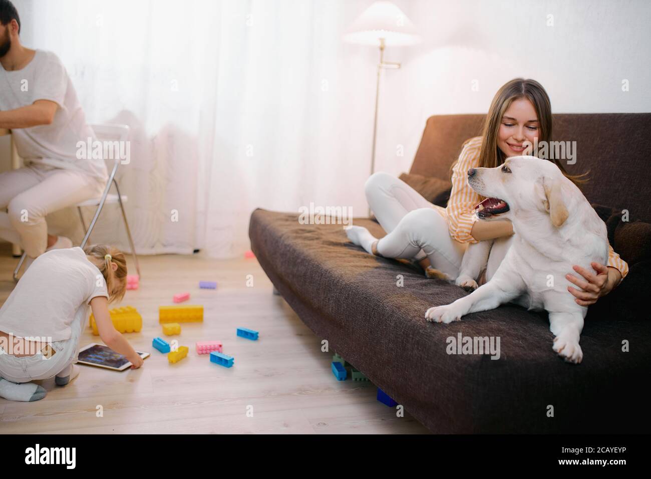 beautiful friendly family spend spare-time at home together, everyone is busy with their own interests, mother playing with pet dog, little daughter w Stock Photo