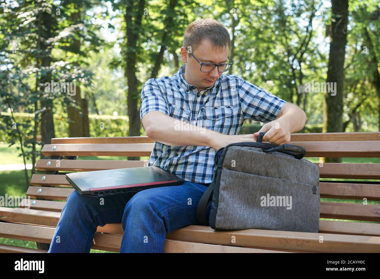 The guy is sitting in the park on a bench. He took out his laptop and looks at things in the bag. Stock Photo