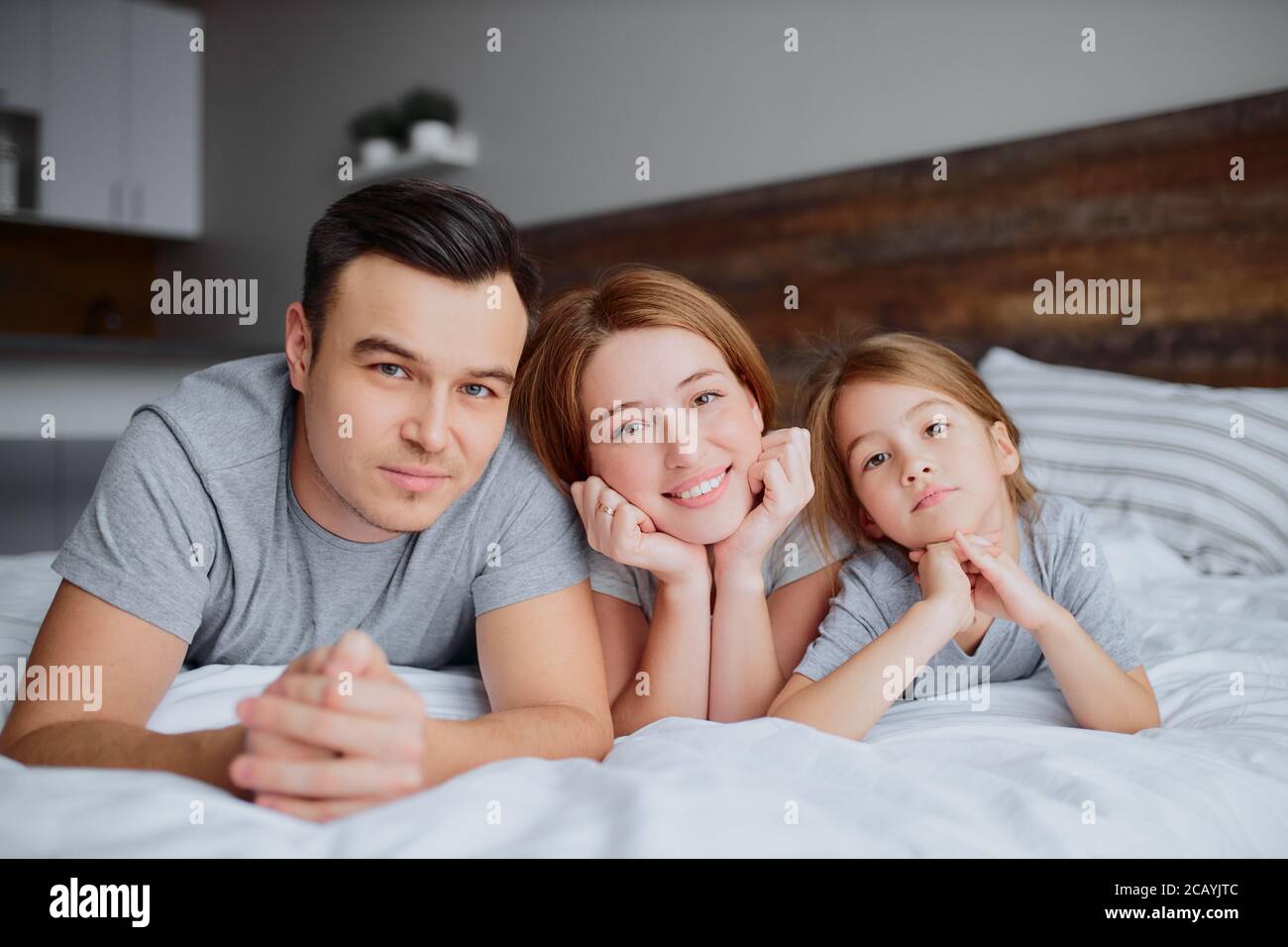 portrait of beautiful happy family lying on bed together, mother father and kid girl kissing hugging and laughing. indoors Stock Photo