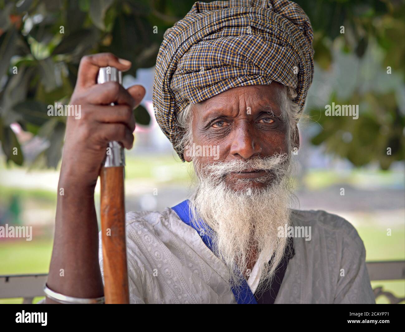 Old Indian Sikh devotee with bloodshot eyes rests on his walking stick and looks into the camera. Stock Photo