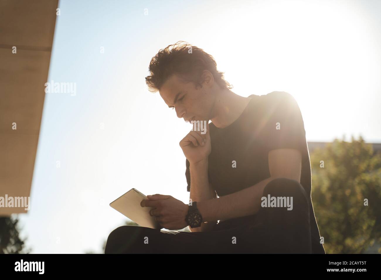 Thoughtful Caucasian young man holding tablet computer while sitting outside university Stock Photo