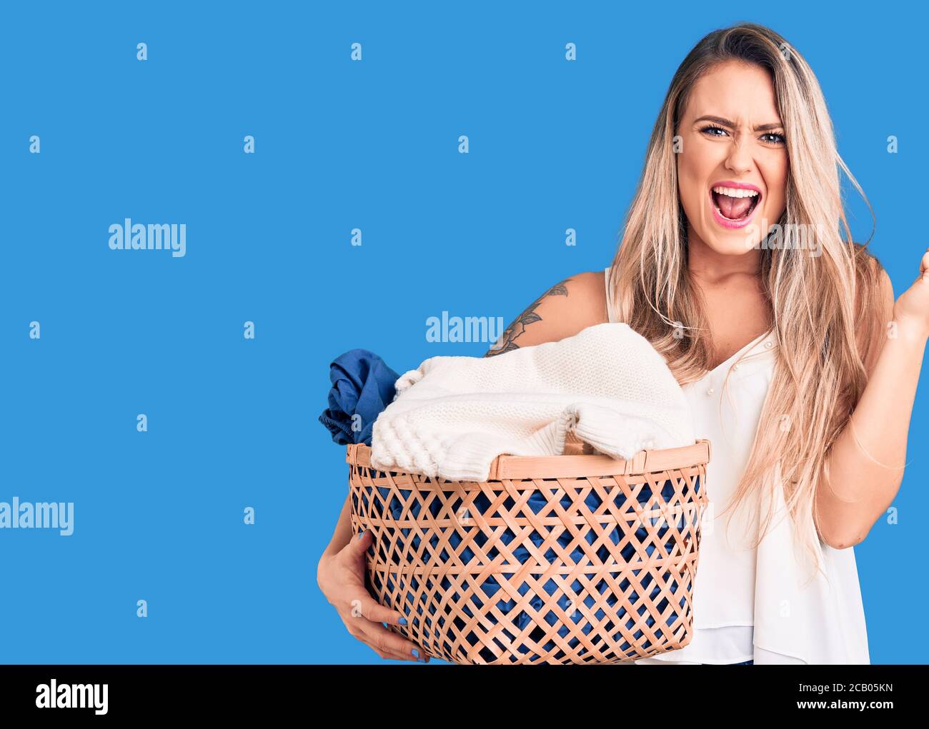 Young beautiful blonde woman holding laundry basket screaming proud ...