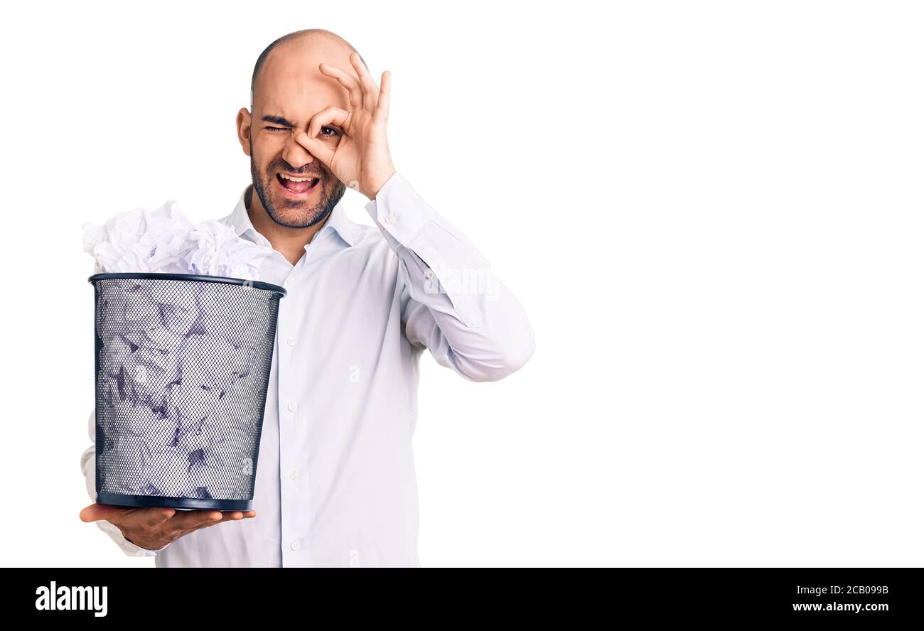 Young handsome man holding paper bin full of crumpled papers smiling happy doing ok sign with hand on eye looking through fingers Stock Photo