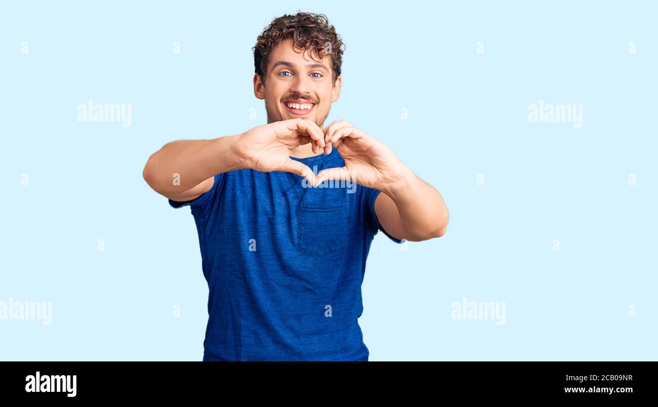 Young handsome man with curly hair wearing casual clothes smiling in love doing heart symbol shape with hands. romantic concept. Stock Photo