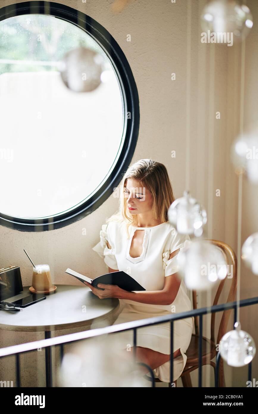 Beautiful young woman sitting at small table in coffeeshop and reading ...