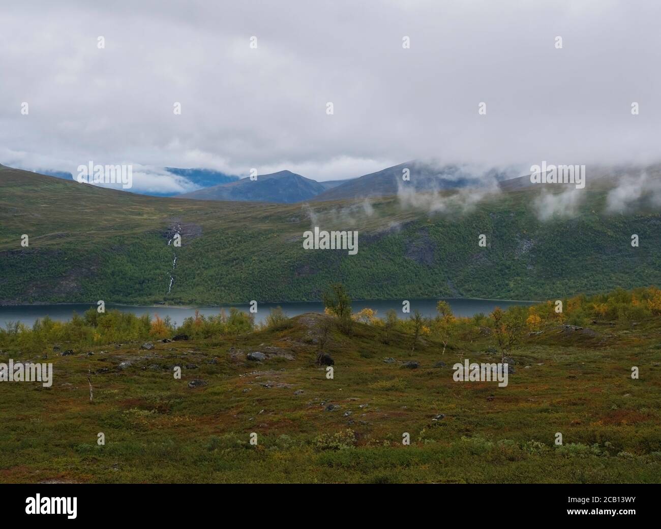 Lapland nature at Kungsleden hiking trail with green mountains, Teusajaure lake, rock boulders, autumn colored bushes, birch tree and heath in mist Stock Photo