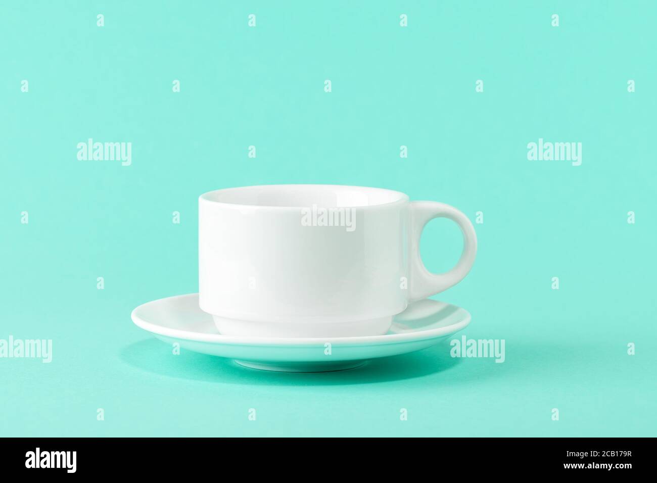 Empty porcelain coffee cup and a saucer over a mint green color background. White ceramic teacup. Modern stylish tableware. Copy space. Front view. Stock Photo