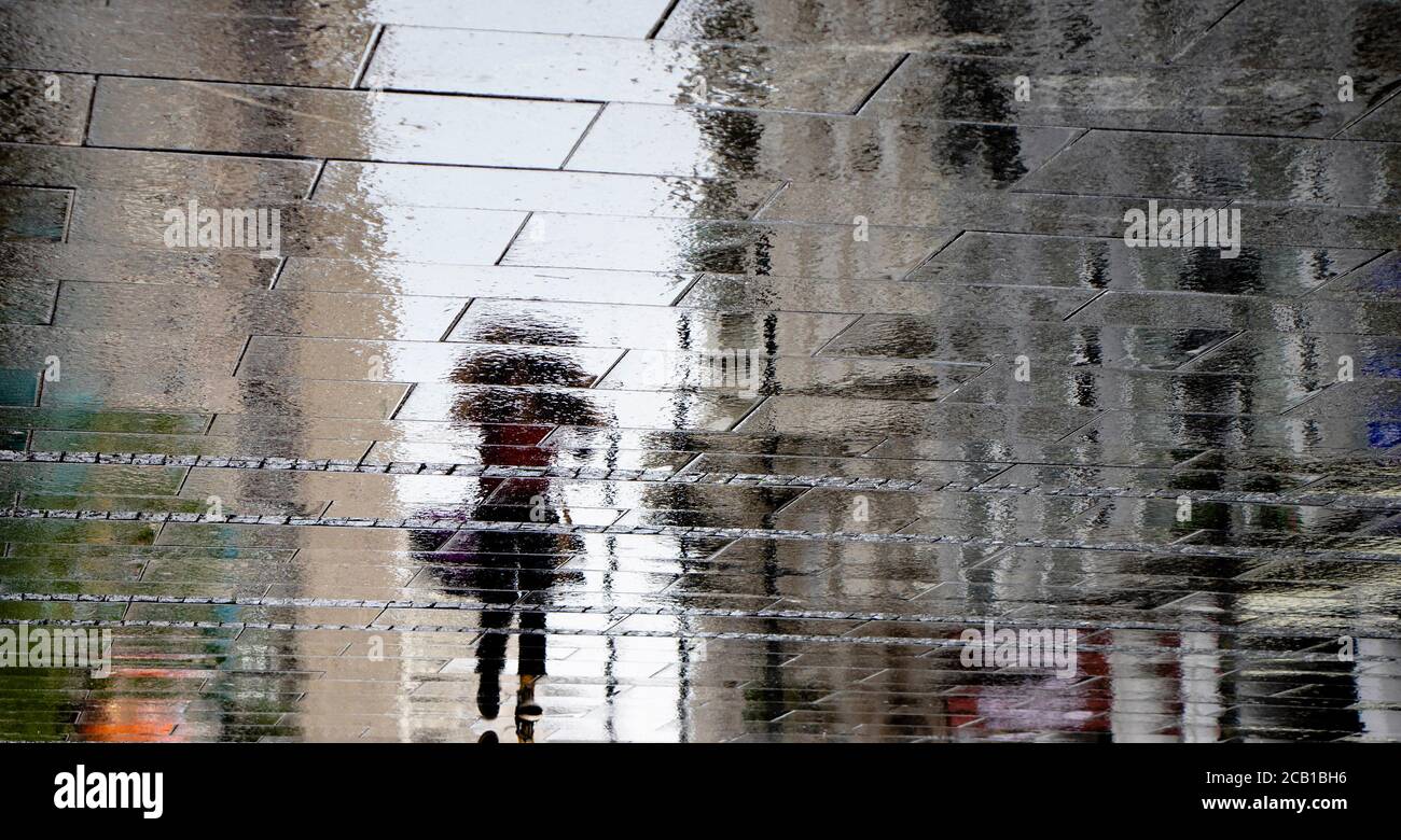 Blurry reflection shadow silhouette on wet puddle of a woman walking with jacket over her had on a city street on a rainy day, abstract background Stock Photo