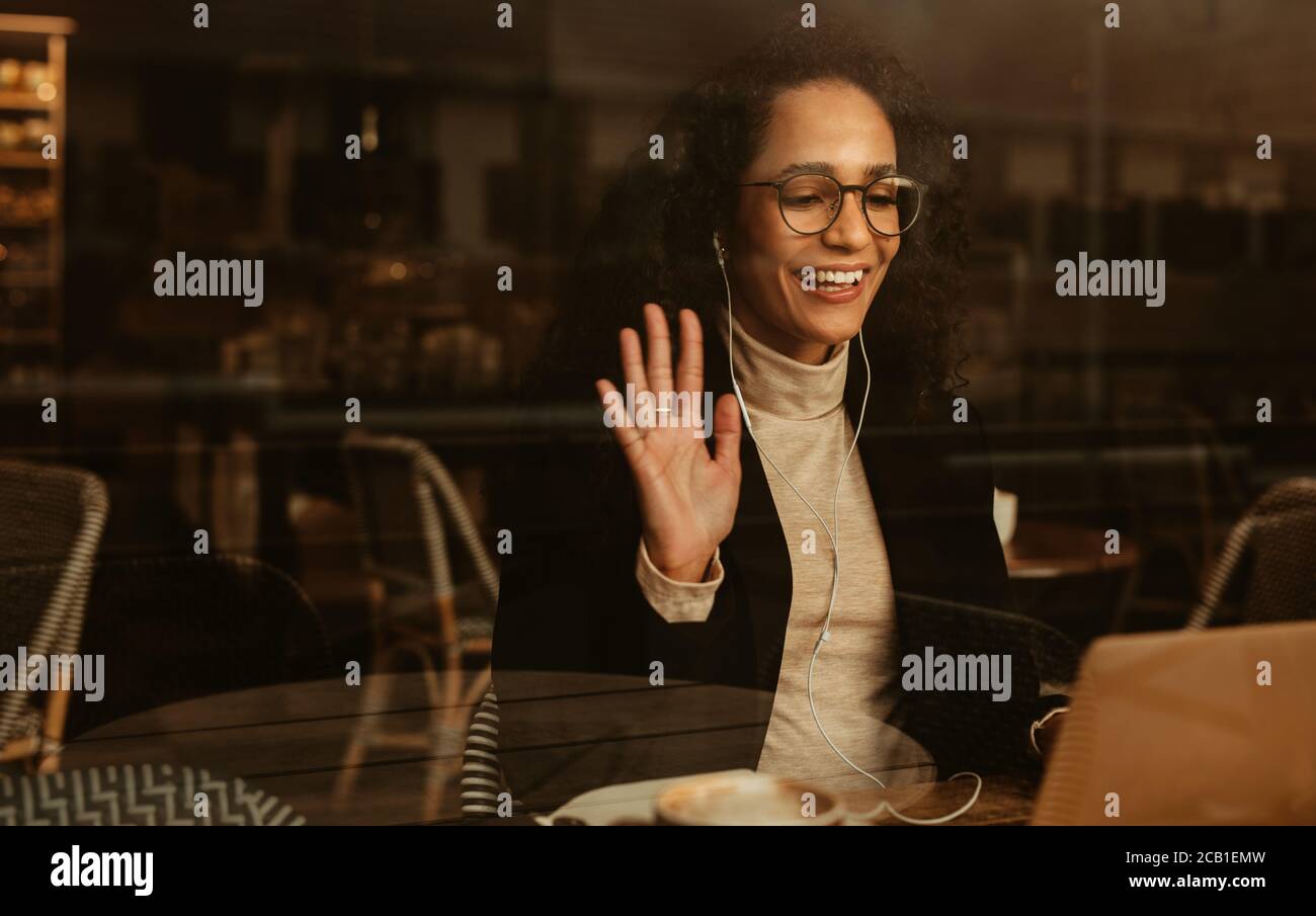 Businesswoman having a video call on her laptop computer. Female professional sitting at cafe wearing earphones waving at the laptop. Stock Photo