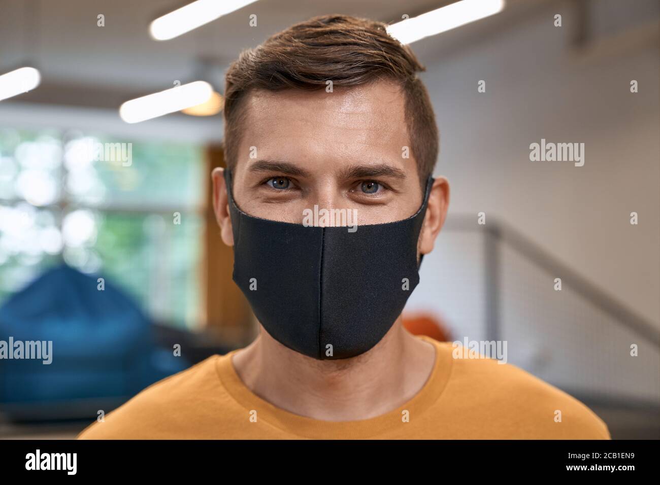 Back to office. Close up portrait of young man, male office worker wearing black protective mask and looking at camera, standing in the modern office Stock Photo