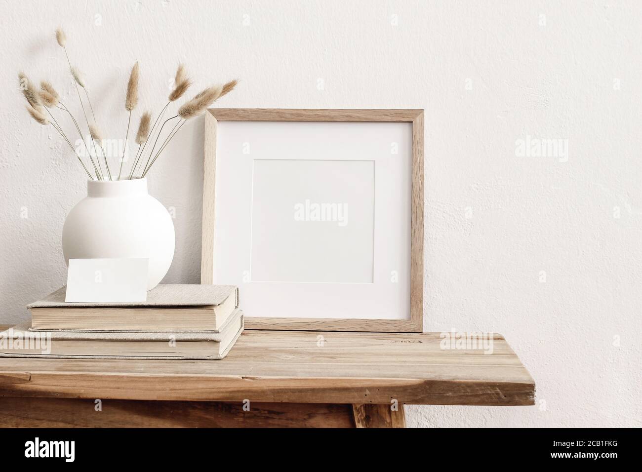 Square wooden frame mockup on vintage bench, table. Modern white ceramic vase with dry Lagurus ovatus grass, books and busines card. White wall Stock Photo