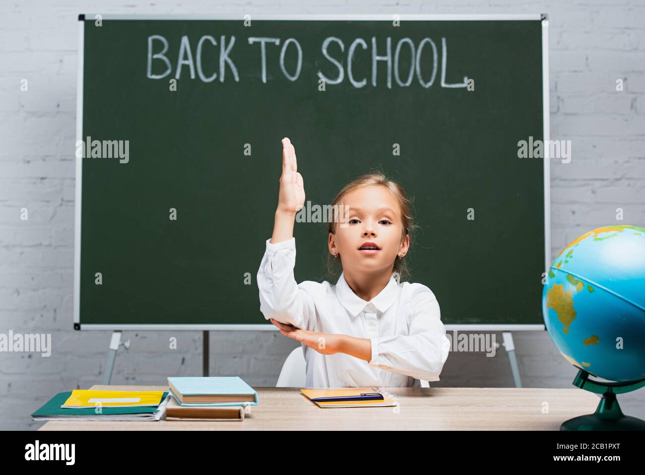 adorable schoolgirl raising hand while sitting at desk near globe and ...