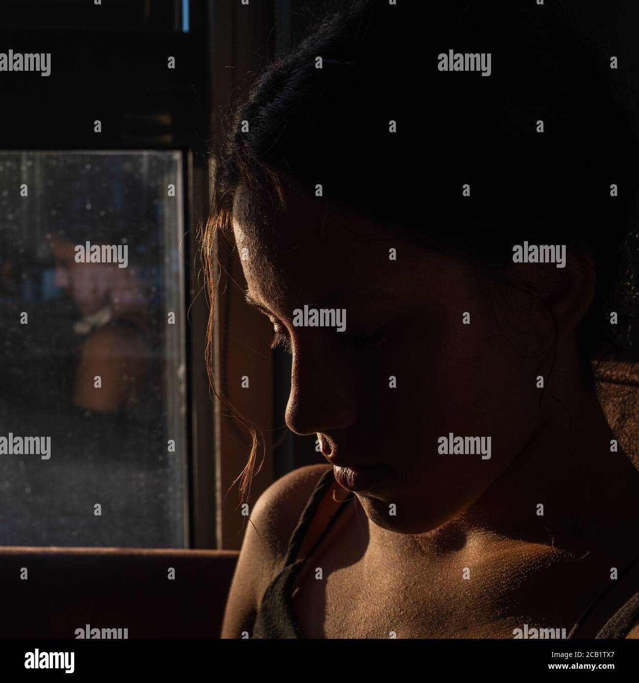 Portrait of beautiful brunette girl in darkness with soft light on her face, pensive silhouette looking down, a reflection on the window. Stock Photo