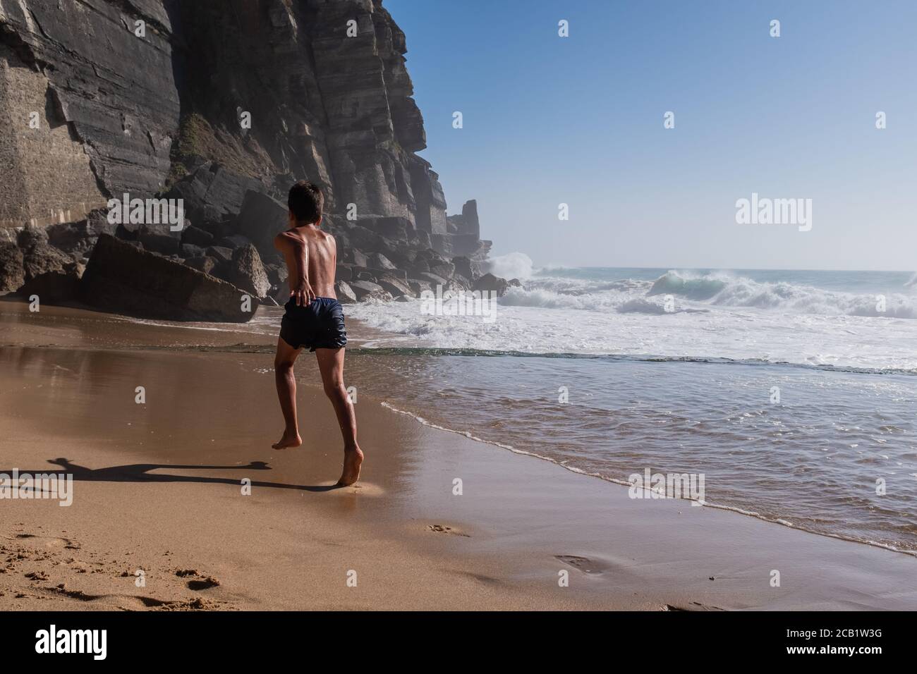 Rear view of boy in blue swim shorts running on sea shore. High cliffs and ocean waves in the background. Active and emotional holiday concept. Stock Photo