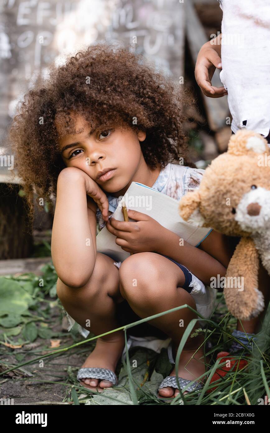 upset and poor african american kid holding notebook near child standing with dirty teddy bear Stock Photo