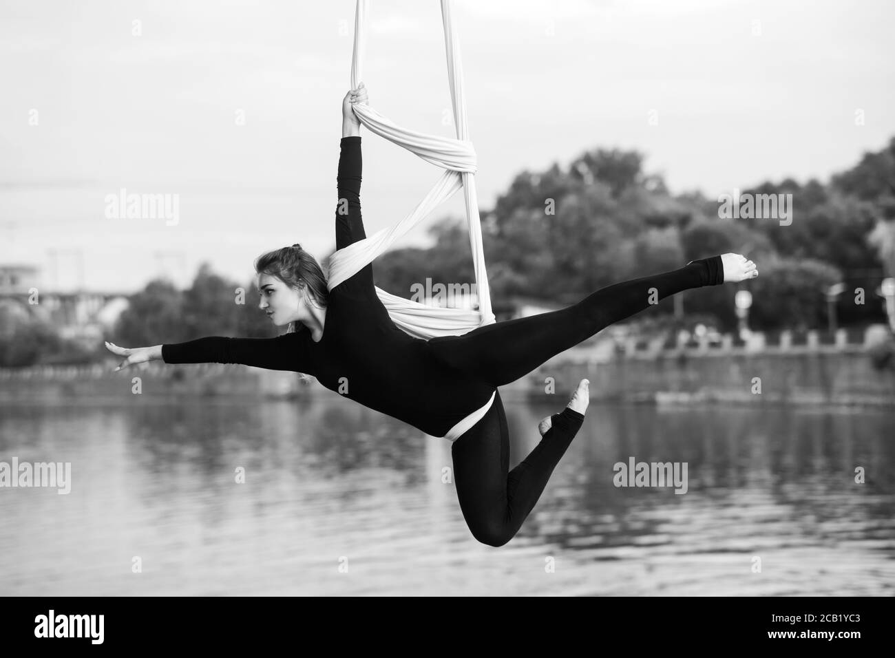 Woman aerialist performs acrobatic tricks on hanging aerial silk against background of river, sky and trees. Black and white image. Stock Photo