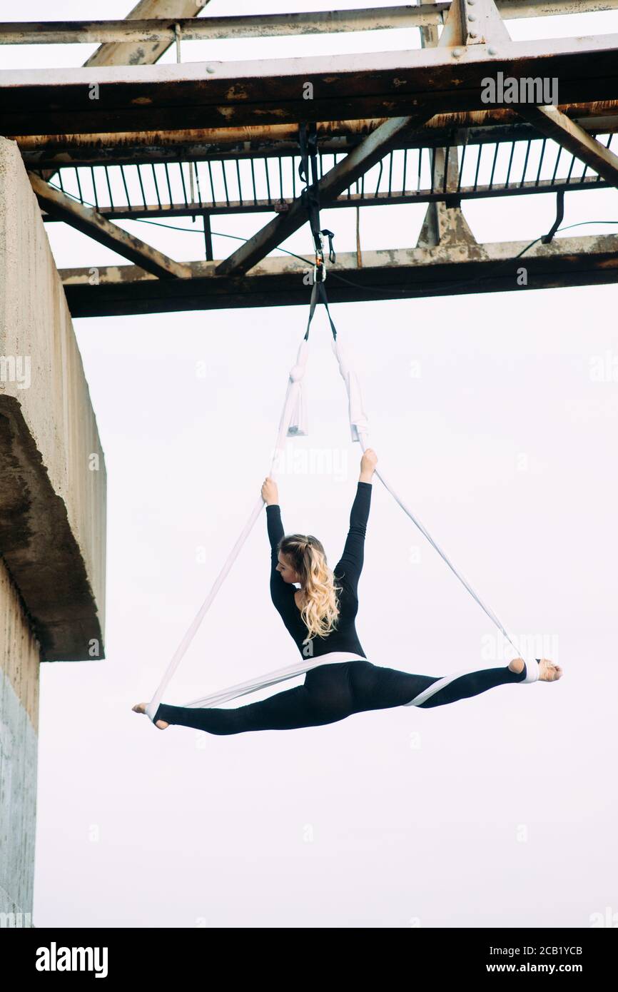 Woman aerialist performs gymnastic split on hanging aerial silk attached to the bridge support against background of sky. Stock Photo