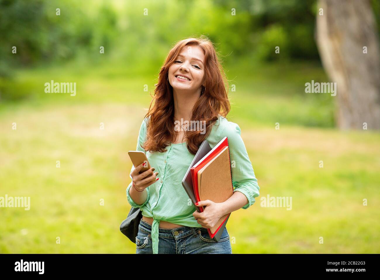 Beautiful Ginger Girl Student Holding Smartphone And Workbooks, Relaxing Outdoors After Classes Stock Photo