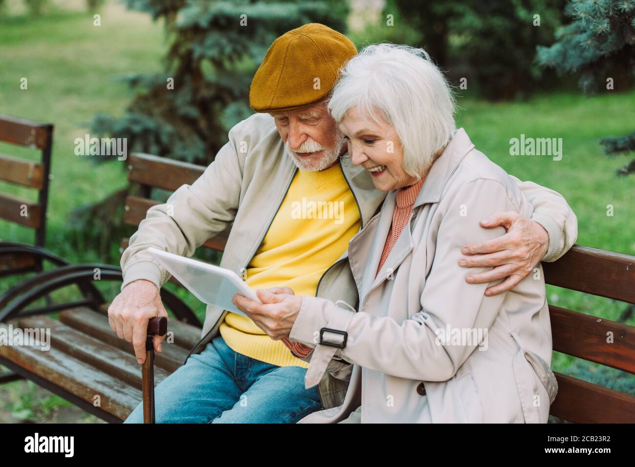 Positive senior woman using digital tablet near husband on bench in ...