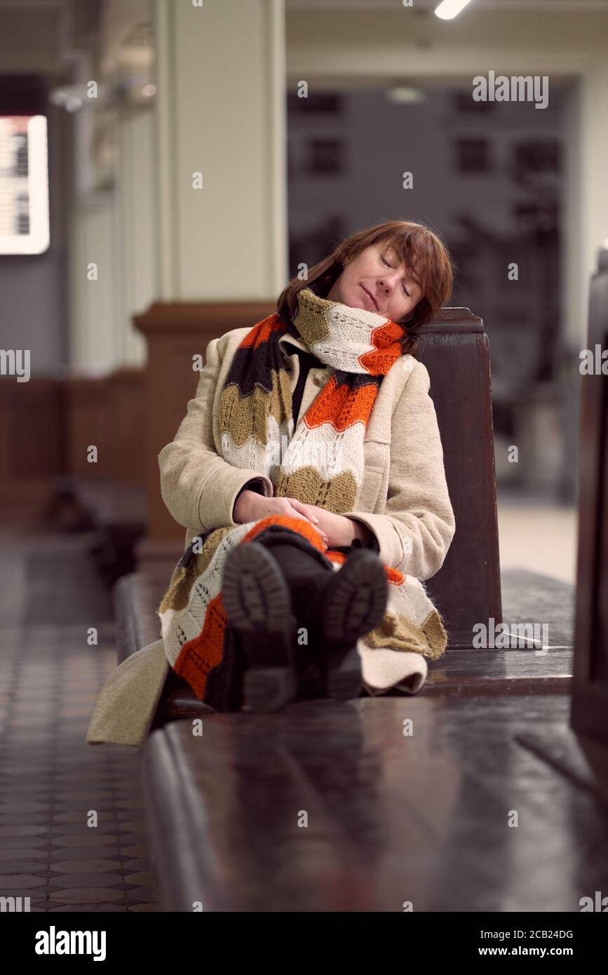Train Station. Mature woman in warm clothes is sitting in waiting room and waiting for train. Evening is dark. Vertical Stock Photo