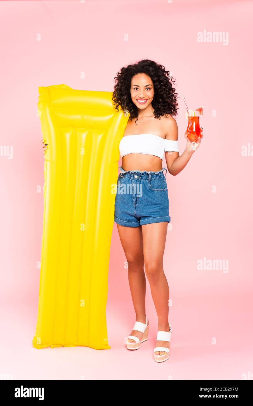 smiling african american girl holding cocktail and inflatable mattress on pink Stock Photo