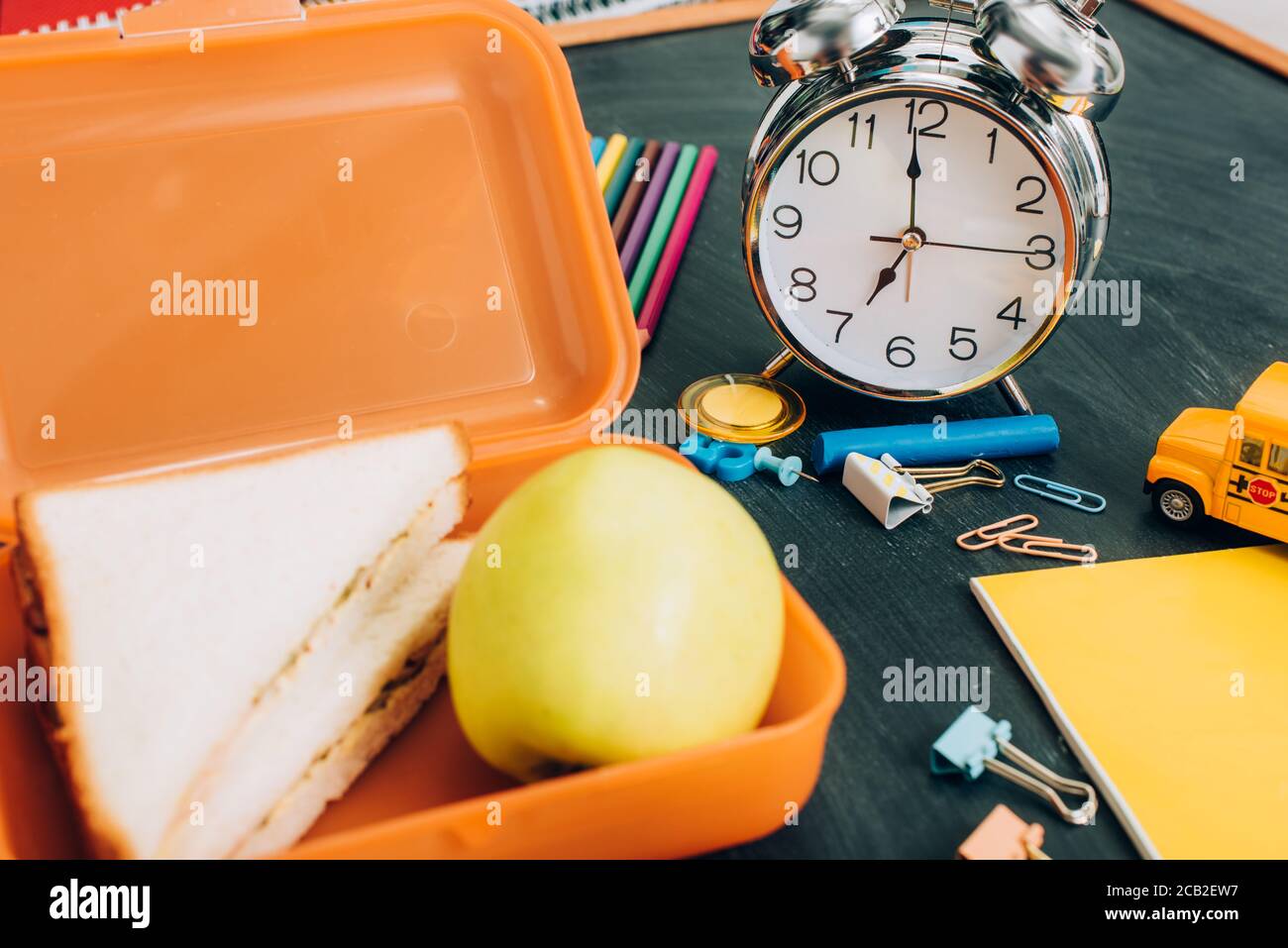 selective focus of lunch box with tasty sandwiches and ripe apple near vintage alarm clock and school supplies on black chalkboard Stock Photo