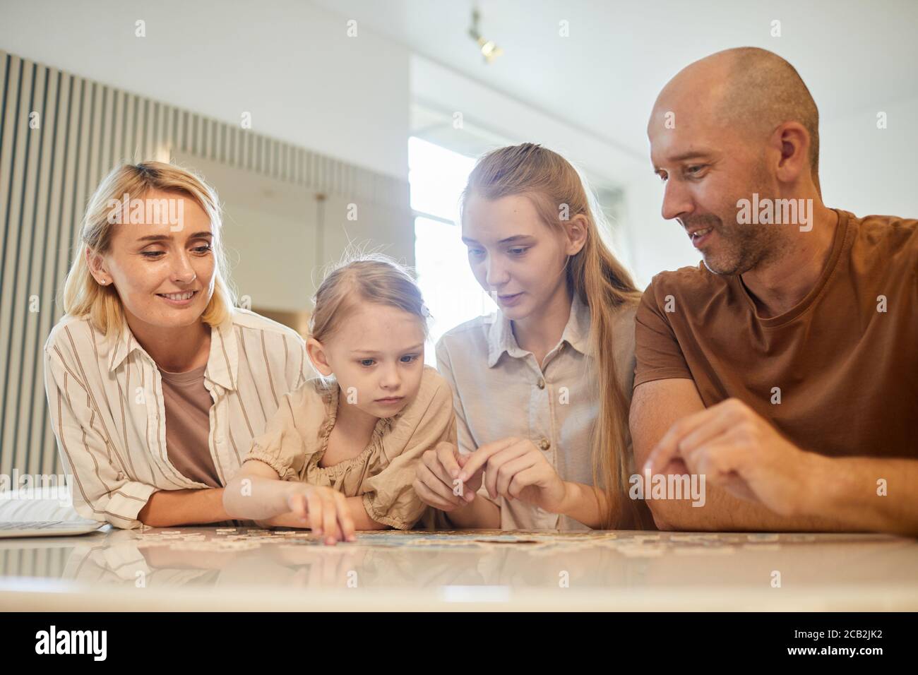 Warm-toned low angle portrait of modern family with two kids solving puzzle together while enjoying time indoors at home, copy space Stock Photo