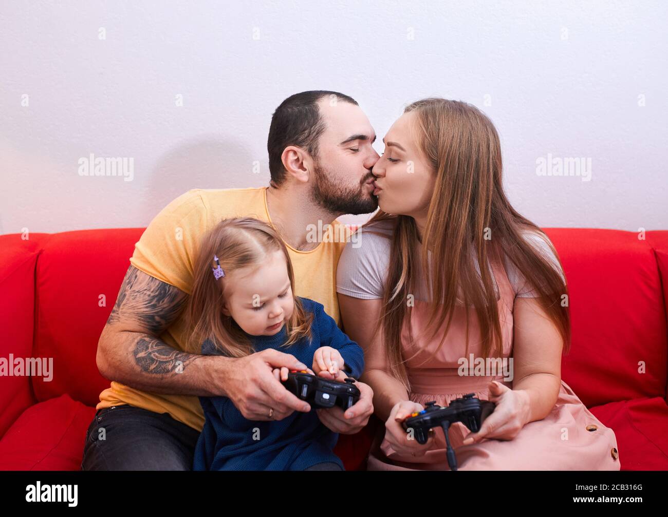 caucasian adorable young parents spend time playing video games with joysticks, sitting on red sofa, modern married couple and cute daughter
