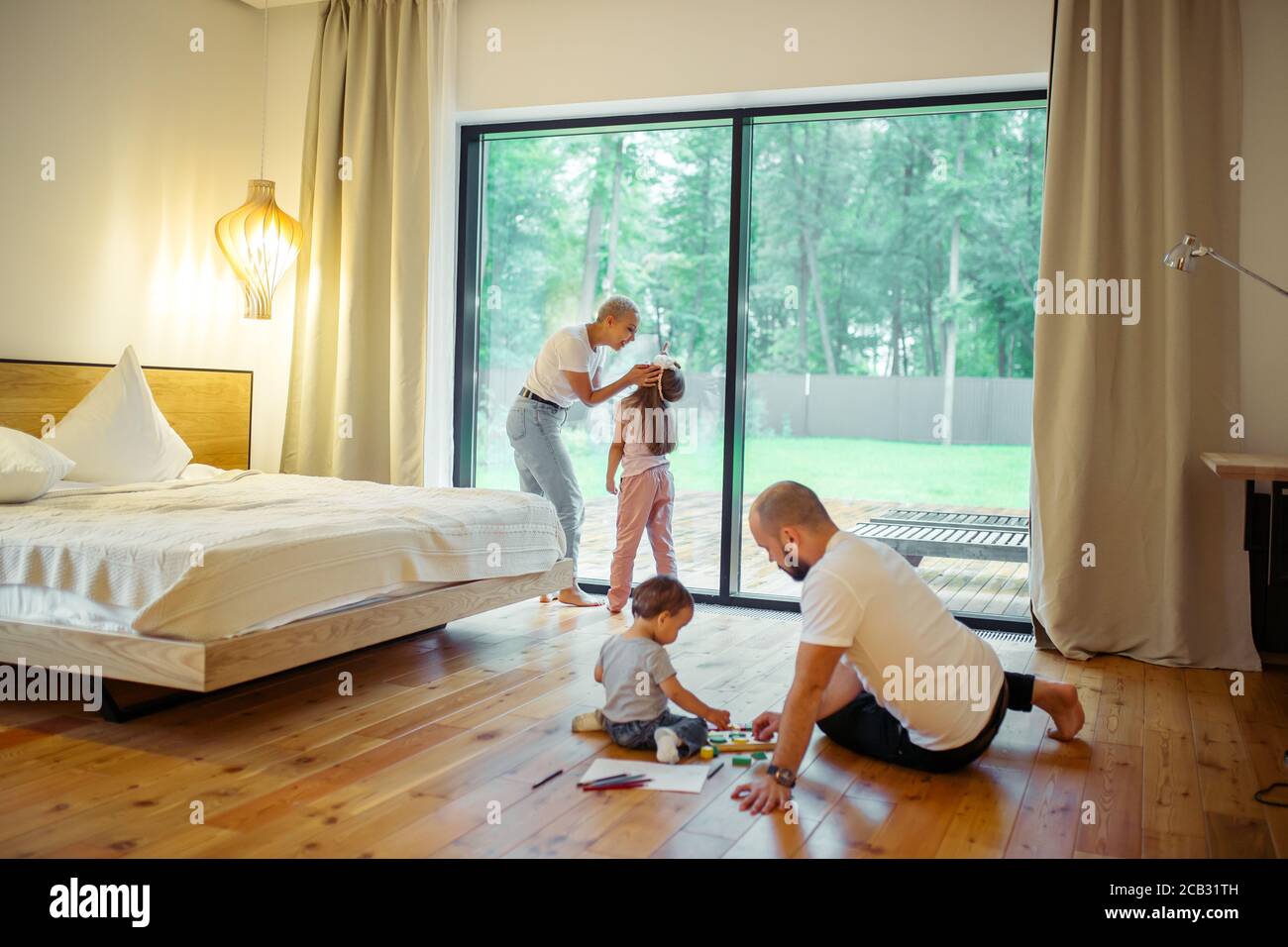 Panoramic window in room. Young parents with their children in room. Mother and daughter, father with son playing on floor. Growing generation, family Stock Photo