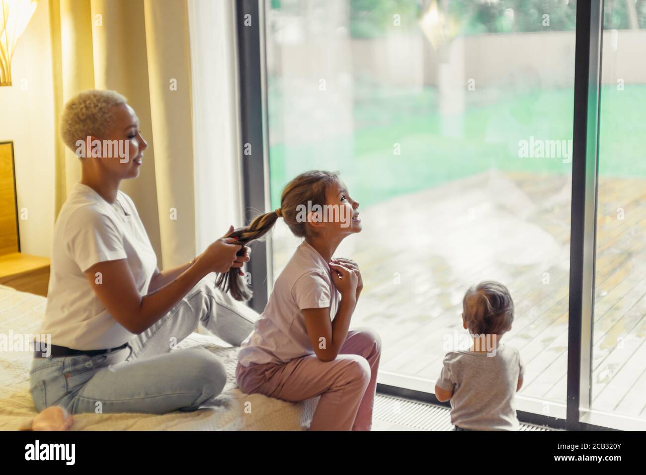 Young woman braid hair to daughter, sitting on bed in weekend. Background panoramic window, baby stand opposite window Stock Photo