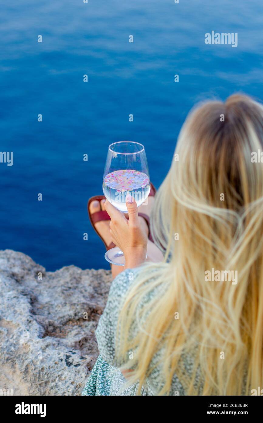 A girl with blond hair sits on the seashore with glass in her hands. Stock Photo