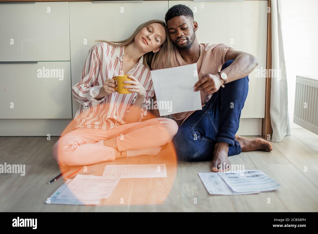 Happy young african husband and his beautiful caucasian blonde wife doing together home budget accounting sitting on kitchen floor with paper sheets a Stock Photo