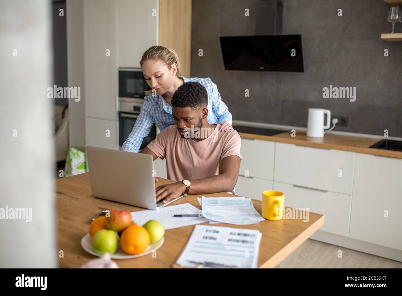 Young mixed race married couple having breakfast at the morning in kitchen and using laptop. Online shopping. Stock Photo