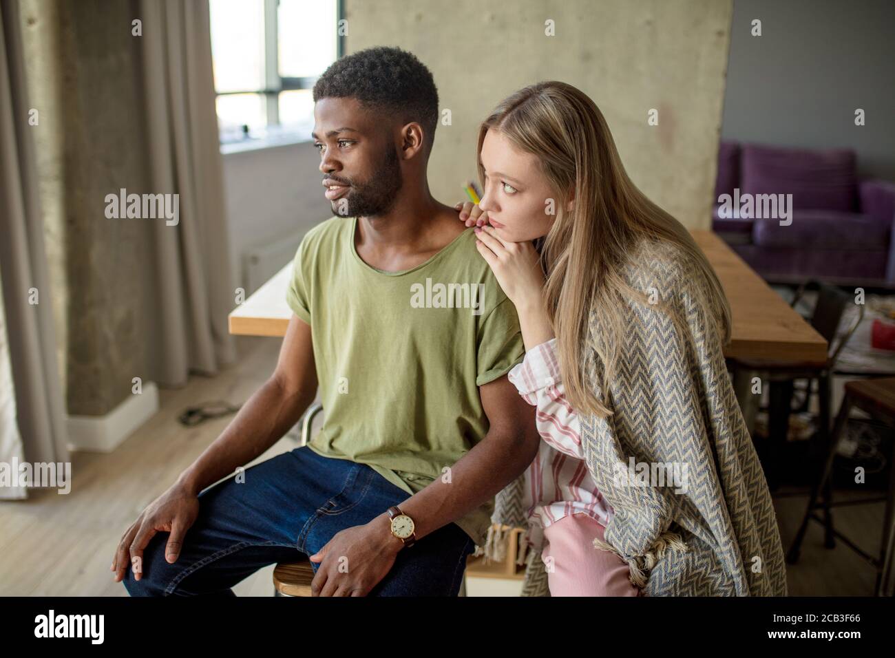 Caucasian blonde woman consoles and supports her african hipster boyfriend, who is having gloomy and frustrated look, couple is sitting in cozy living Stock Photo